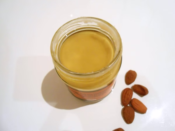 Open jar of Giannetti Artisans Unsweeteend Almond Paste to display the natural almond oil with loose almond nuts around the jar on a white background