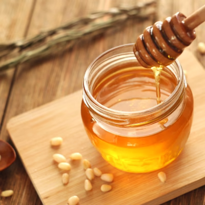 Honey jar on a wooden table with honey flowing down a honey stick into the jar. Nearby are pine nuts and rosemary for Giannetti Artisans Artisan Honey Collection