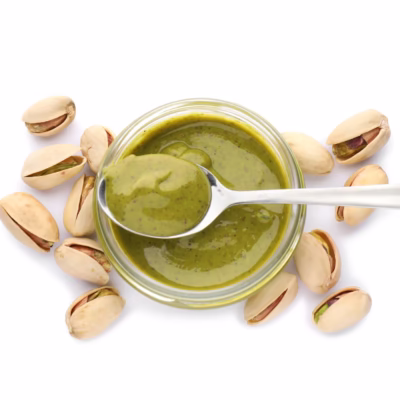 Top view of an open pistachio spread jar with a spoon laying over the jar with pistachios surrounding the jar on a white background for the Giannetti Artisans Artisan Sauces & Spreads Collection
